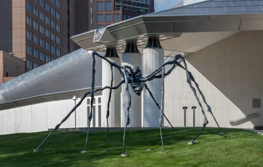 A large dark metal spider sculpture sits on a grassy hill in front of a white modern museum building in the city.
