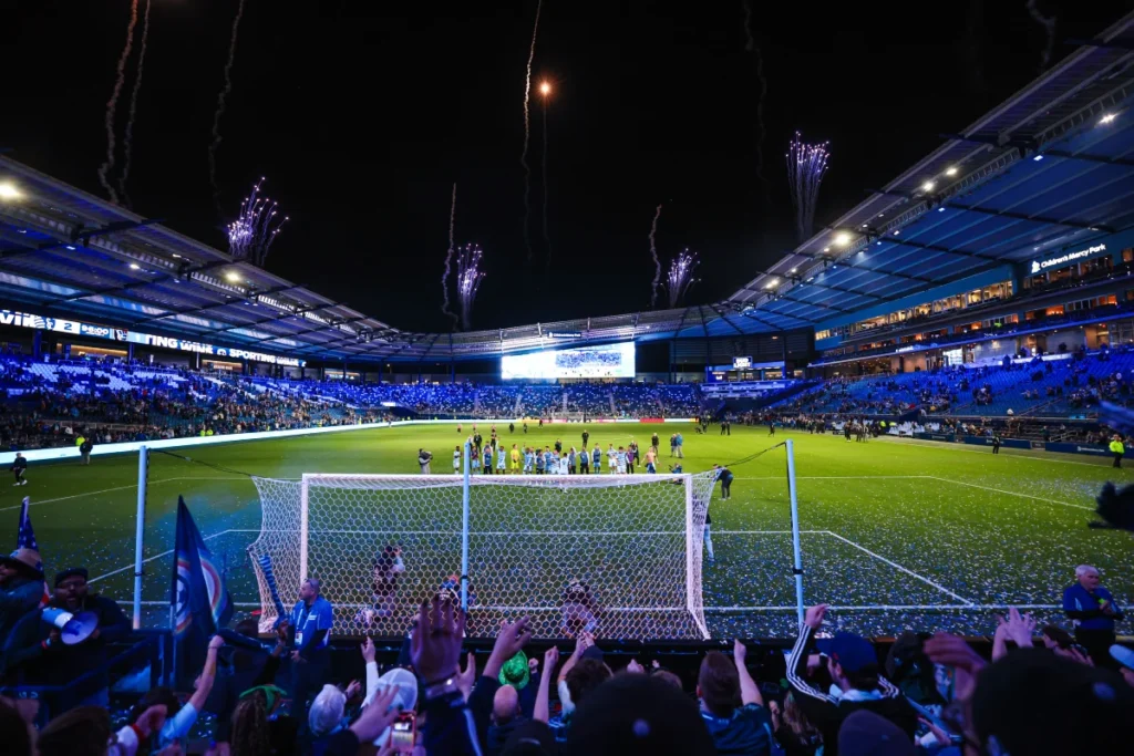 A packed Kansas City soccer stadium hosts a major night event, featuring fans cheering on the field and colorful firewor