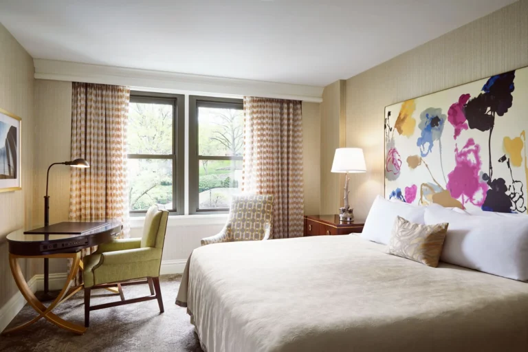 A luxurious hotel room featuring a large bed, colorful abstract art headboard, an elegant wood desk, and windows overloo