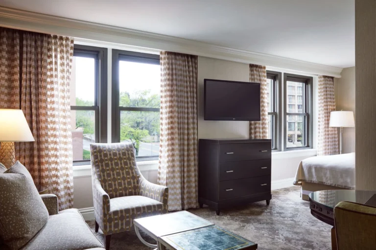 A spacious luxury hotel guest room featuring a patterned armchair, sofa, wall-mounted TV, and large windows with decorat