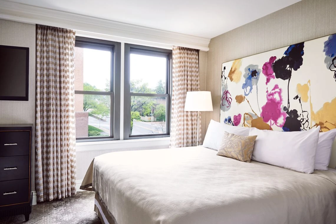 A bright boutique hotel bedroom suite featuring a queen bed, crisp white linen, and a colorful abstract floral headboard