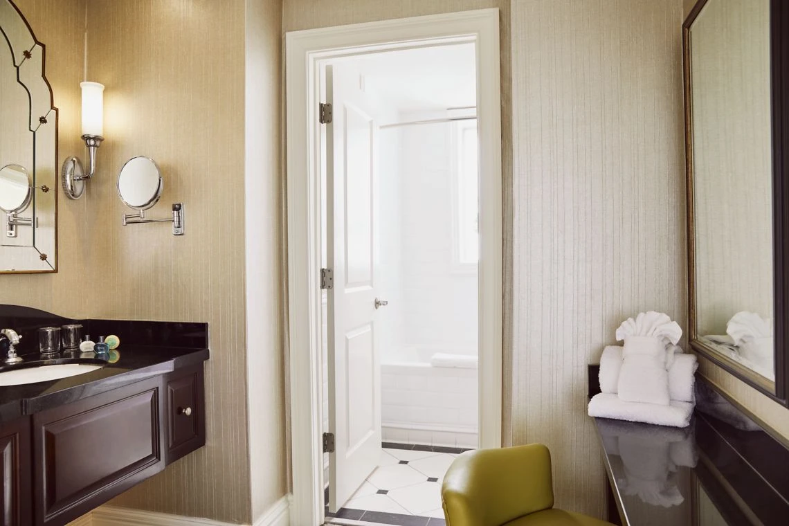 A sophisticated hotel suite bathroom shows a dark vanity counter, plush white folded towels, a green chair, and the door