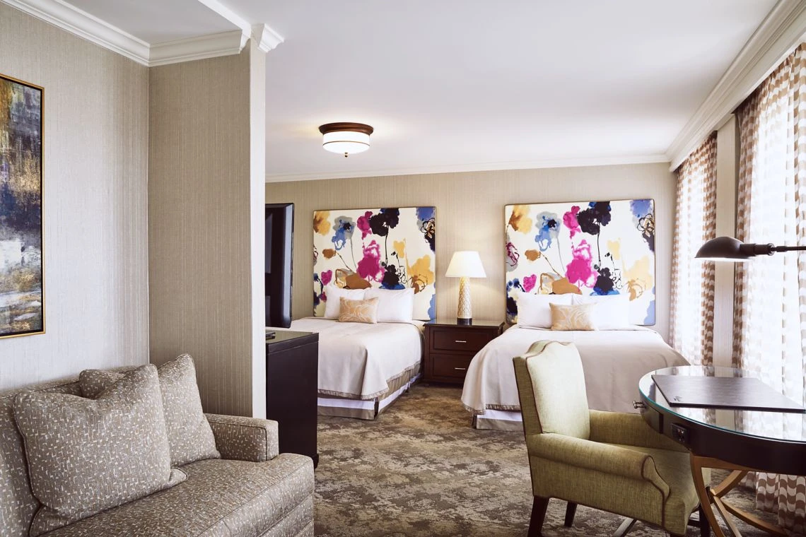A spacious luxury hotel suite featuring two beds with vibrant floral headboards, a separate sitting area with a couch, a