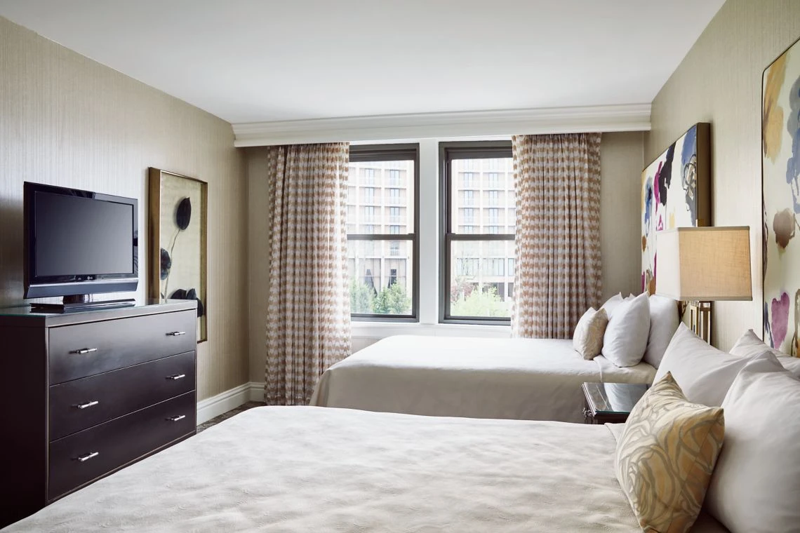 A modern, luxury hotel suite in Kansas City featuring two double beds, patterned curtains, a dresser, TV, and abstract w
