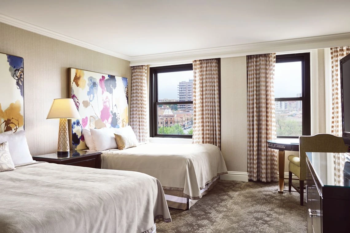 A brightly lit luxury hotel room featuring two double beds, abstract floral artwork, and a large window overlooking a ci