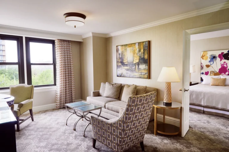 A luxury hotel suite living room features neutral seating, large windows, and an open doorway to the spacious, modern be