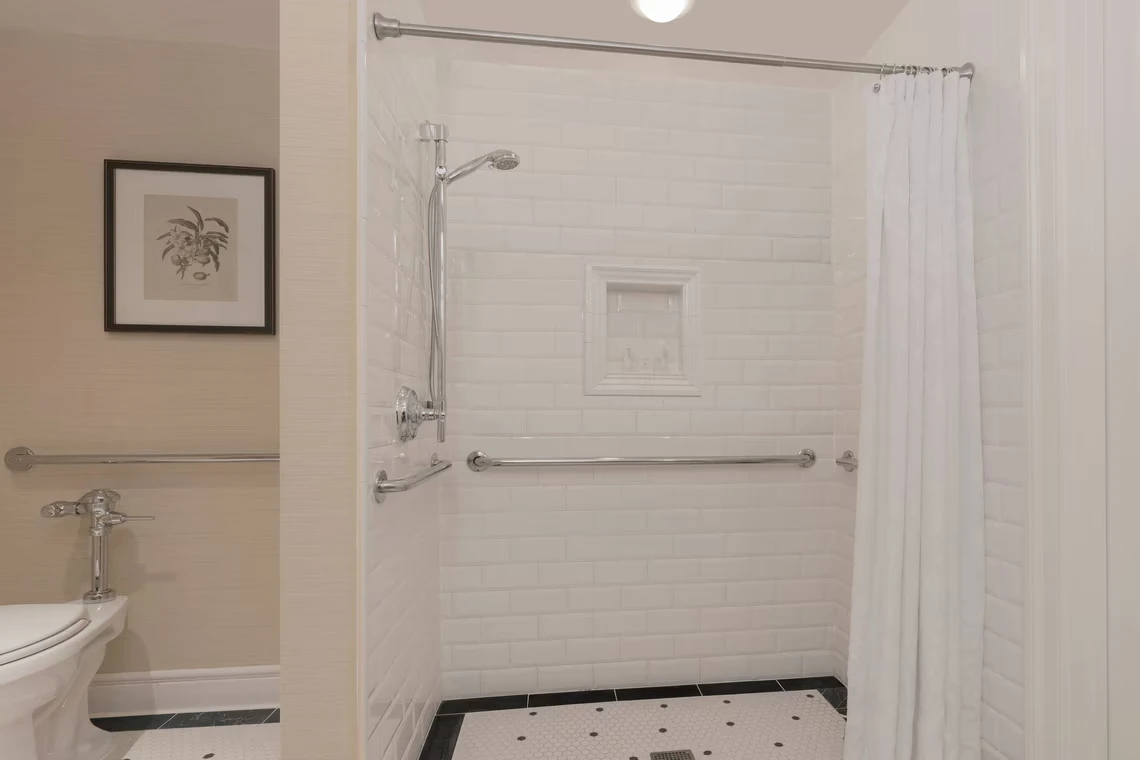 A bright, accessible hotel shower area featuring white subway tiles, chrome grab bars, a handheld shower, and classic mo