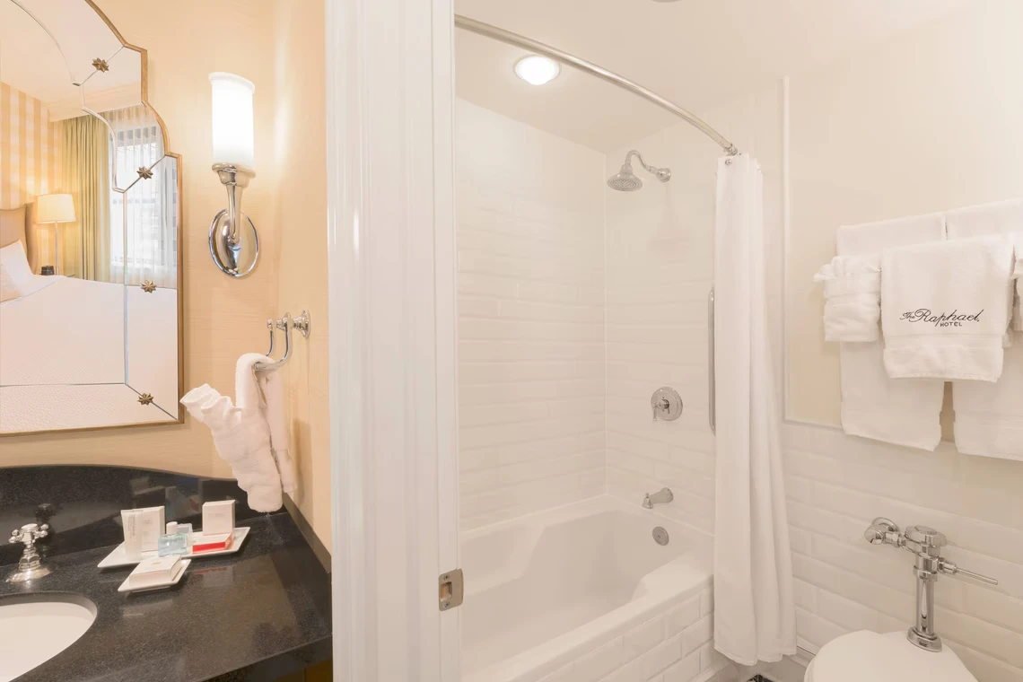 A bright boutique hotel bathroom featuring a white tile shower-tub, dark granite vanity, modern fixtures, and luxurious