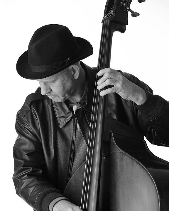 A black and white photo of a musician wearing a fedora and leather jacket playing an upright bass, highlighting the Kans