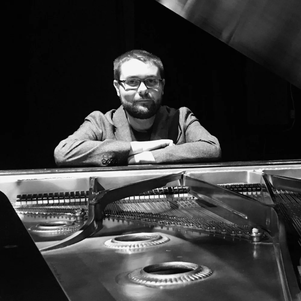 Black and white image of a musician in glasses leaning on a grand piano, suggesting a live jazz performance setting.