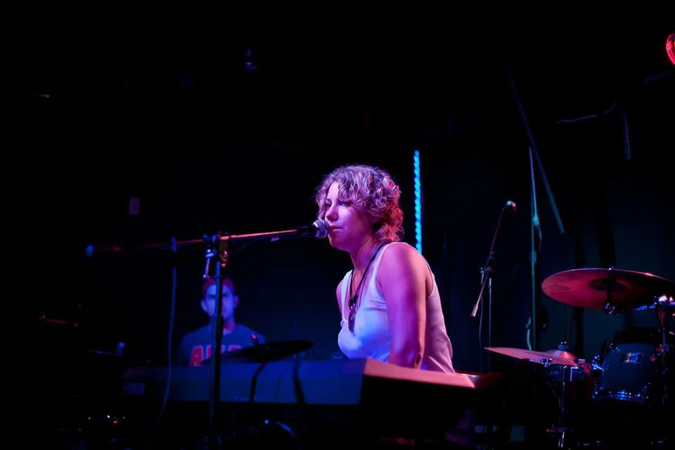 Female musician singing and playing the keyboard illuminated by magenta and blue stage lights during a live music perfor