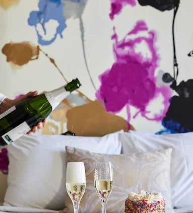 A hand pours champagne into a glass next to a colorful birthday cake on a plush bed, emphasizing a romantic luxury hotel