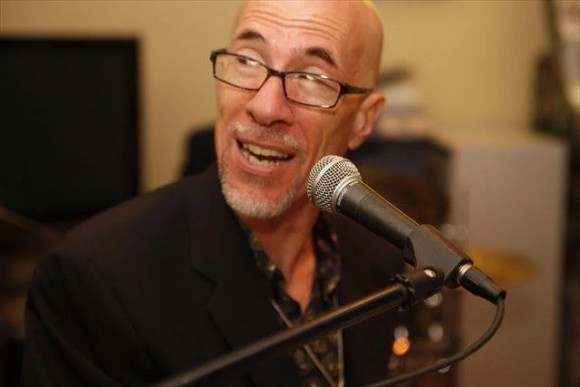 Close-up of a smiling male musician with glasses performing live jazz vocals into a microphone in a dimly lit venue.