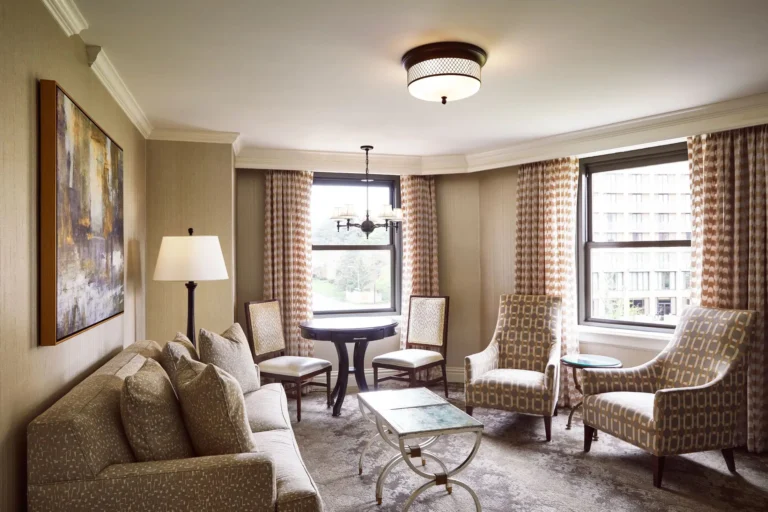 An elegantly decorated luxury hotel suite sitting area features a sofa, patterned armchairs, and large windows with orna