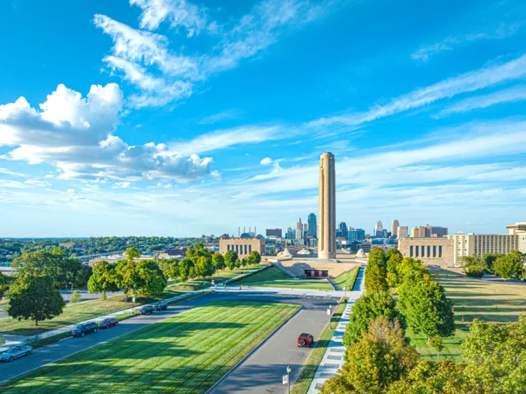 An aerial view of a tall stone monument rising above green park grounds, flanked by trees and a roadway, with a modern c