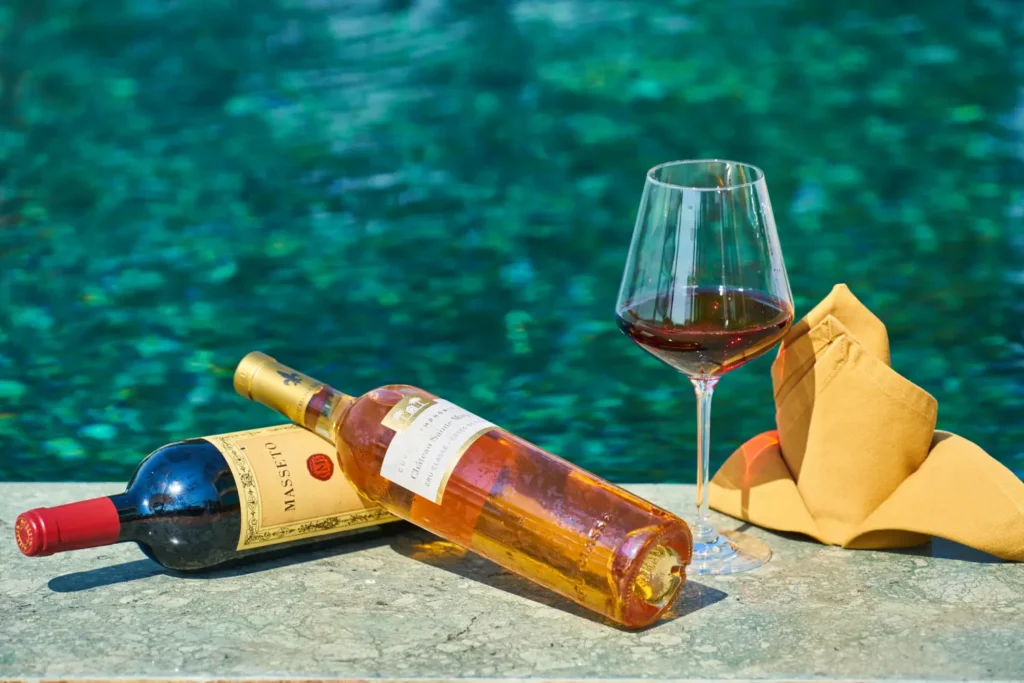 A chilled bottle of rosé wine and a glass of red wine sitting on the edge of a sparkling blue hotel pool, suggesting a l