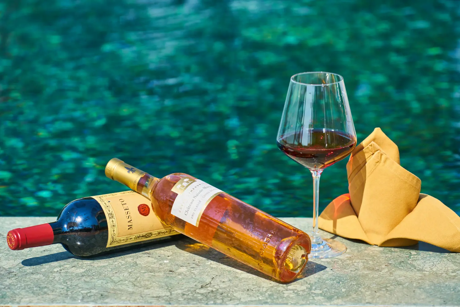 A chilled bottle of rosé wine and a glass of red wine sitting on the edge of a sparkling blue hotel pool, suggesting a l