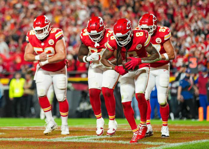 Four football players in red jerseys run down the field during a night game as fans cheer loudly in the stadium stands.