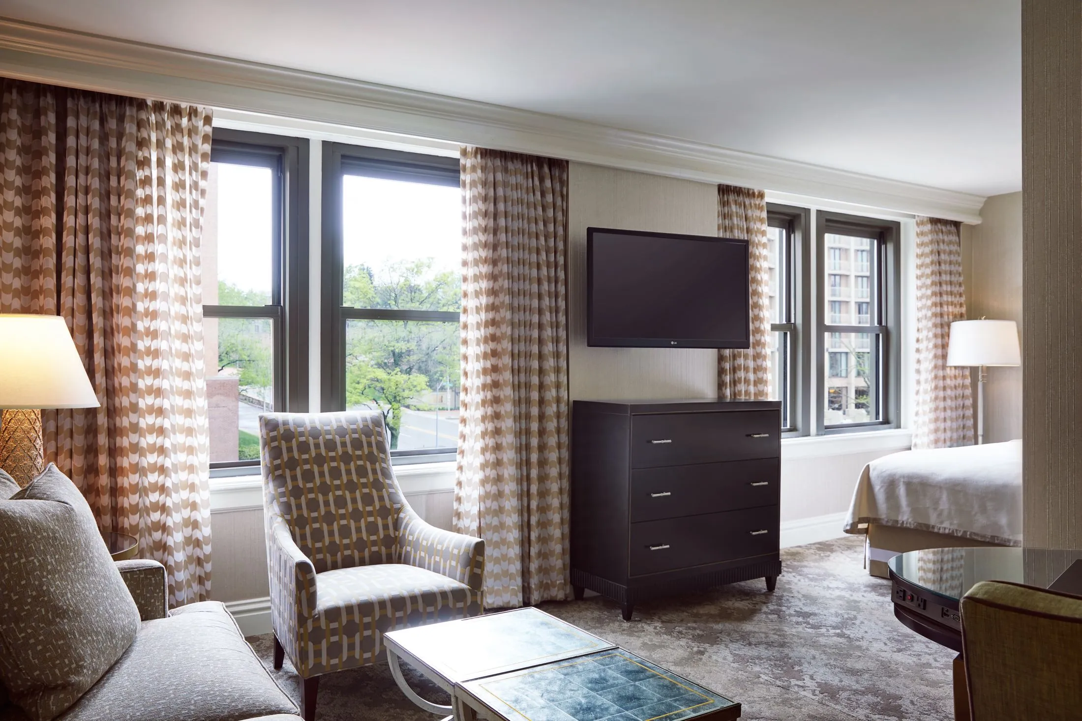 A spacious luxury hotel guest room featuring a patterned armchair, sofa, wall-mounted TV, and large windows.