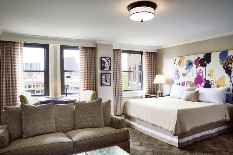 Boutique hotel suite at The Raphael Hotel, featuring a plush taupe couch, king bed with floral art, and natural light.