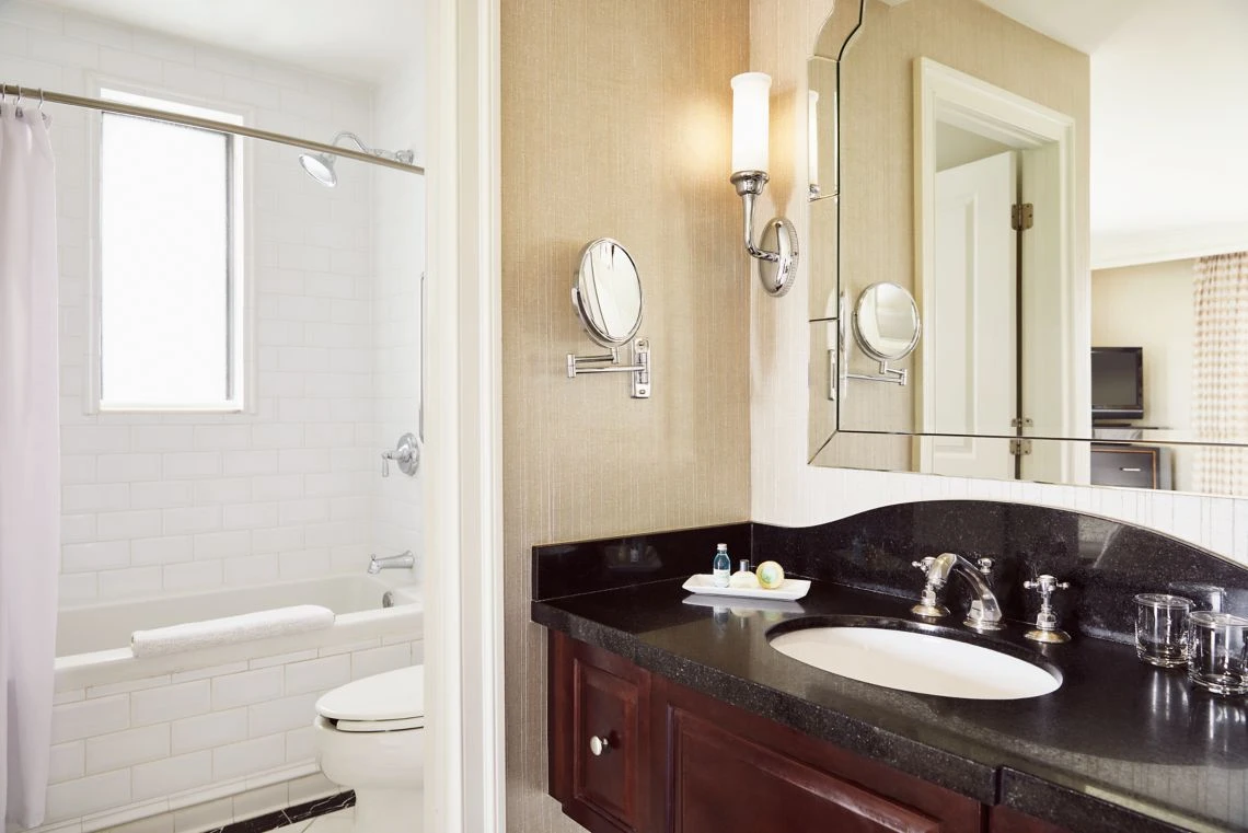 A luxury hotel suite bathroom featuring a dark granite vanity counter, elegant mirror, magnifying glass, and white subwa