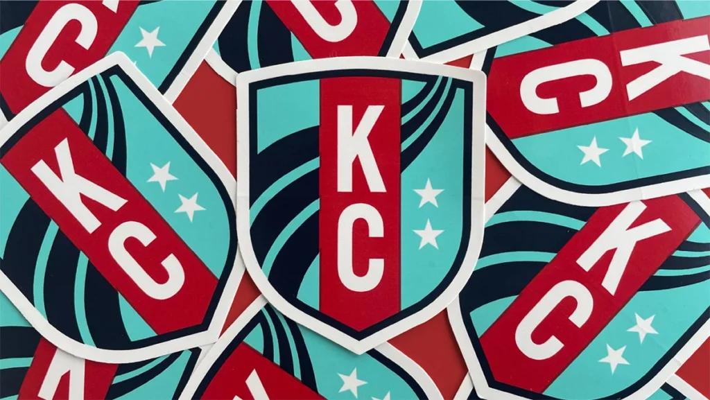 A close-up view of overlapping stylized Kansas City shield logos in teal, red, and navy blue, suggesting local pride and
