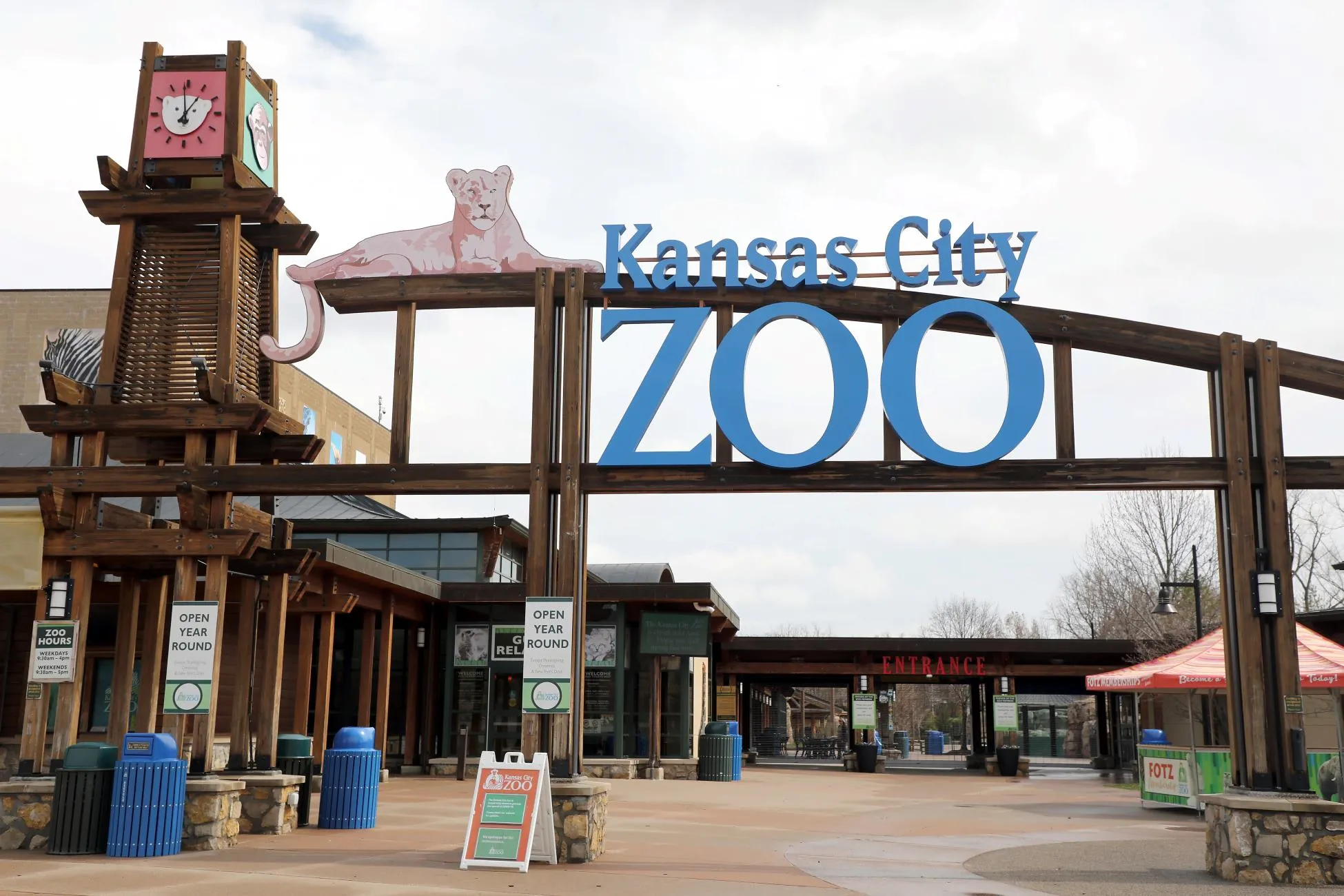 A wide view of the Kansas City Zoo main entrance gate, showing the large wooden structure, a lioness cutout, and the pro