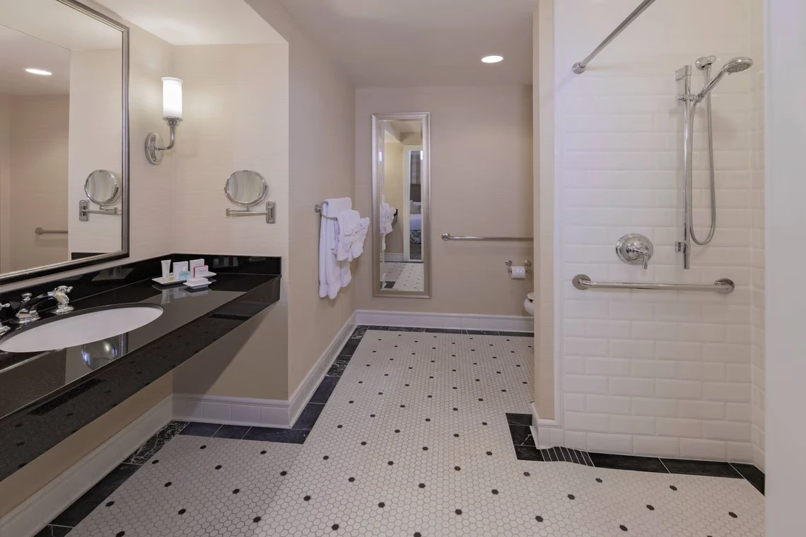 A spacious, upscale hotel bathroom featuring a black granite vanity, classic hexagon tile flooring, and an accessible su