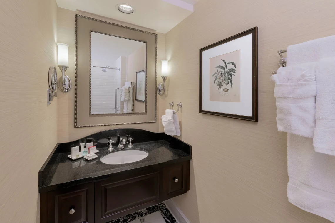 View of an elegant hotel bathroom vanity area with dark cabinetry, toiletries, and framed art on textured beige walls.