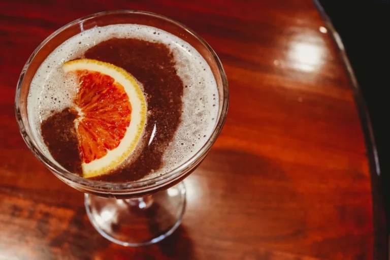 A close-up view of a dark specialty cocktail with foam, garnished with a blood orange slice on a glossy wooden table.