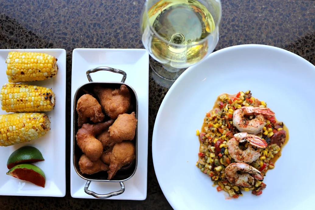 Overhead view of a gourmet seafood dinner with plated shrimp, corn succotash, fried hushpuppies, and grilled corn.
