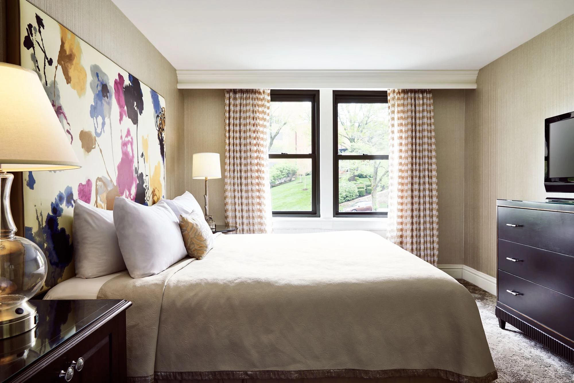 A luxurious hotel bedroom featuring a plush bed, oversized colorful floral headboard, dark wood furniture, and windows w
